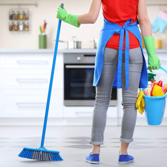 Person ready to clean kitchen with broom and cleaning supplies bucket, wearing red shirt and blue apron.